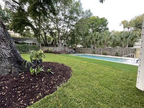 A backyard with a swimming pool, a wooden fence, and a variety of trees and plants.