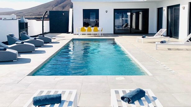 Un área de piscina en la azotea con sillones de descanso y una mesa, una montaña al fondo y un edificio moderno con ventanas amplias.