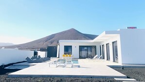 Un edificio moderno con zona de piscina, asientos exteriores y una montaña al fondo.