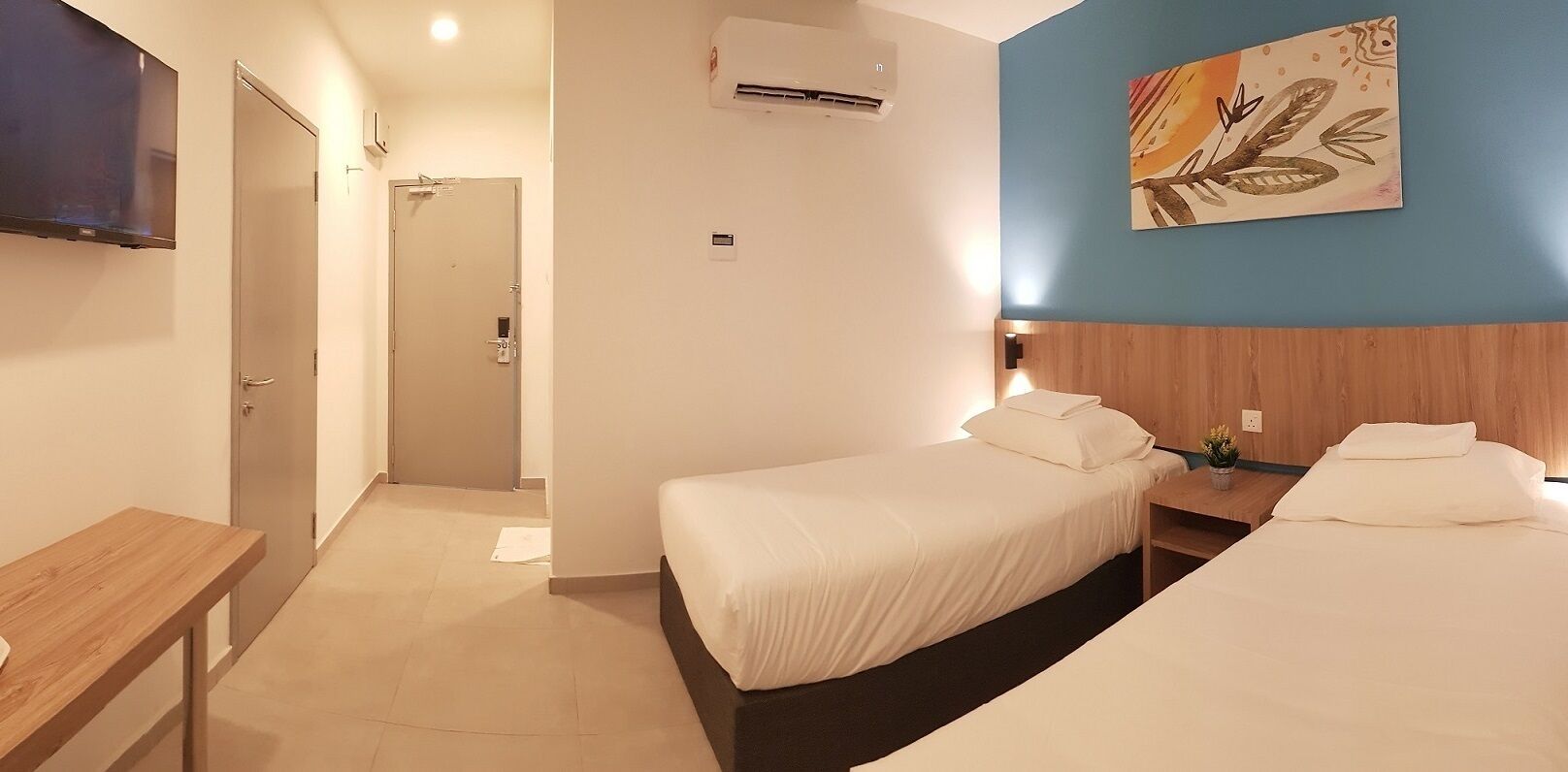 Ein Hotelzimmer mit zwei Betten, einem Holztisch, einem Fernseher, einer Klimaanlage und einem Gemälde an der Wand.