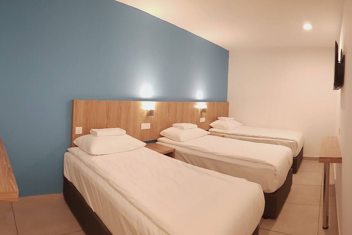 A hotel room with three single beds, a wooden headboard, a blue accent wall, and a small table with a lamp.