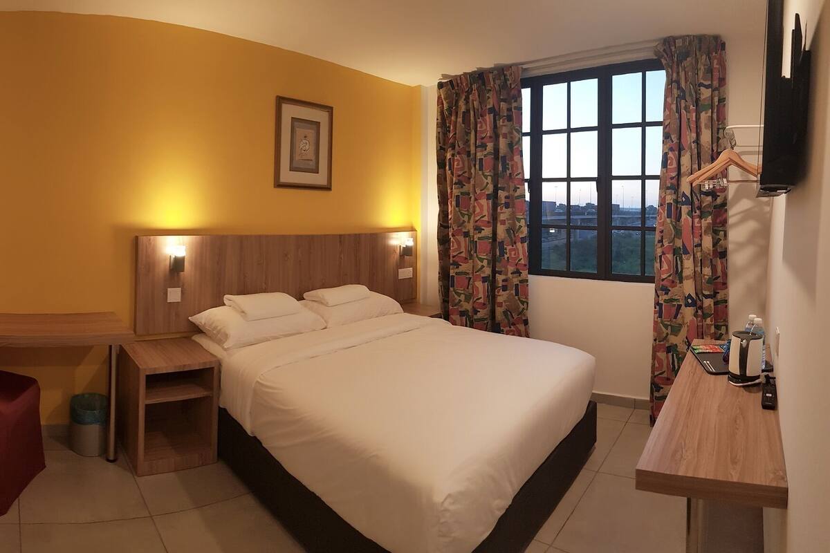A hotel room with a bed, a desk, a chair, a TV, and a window with curtains.