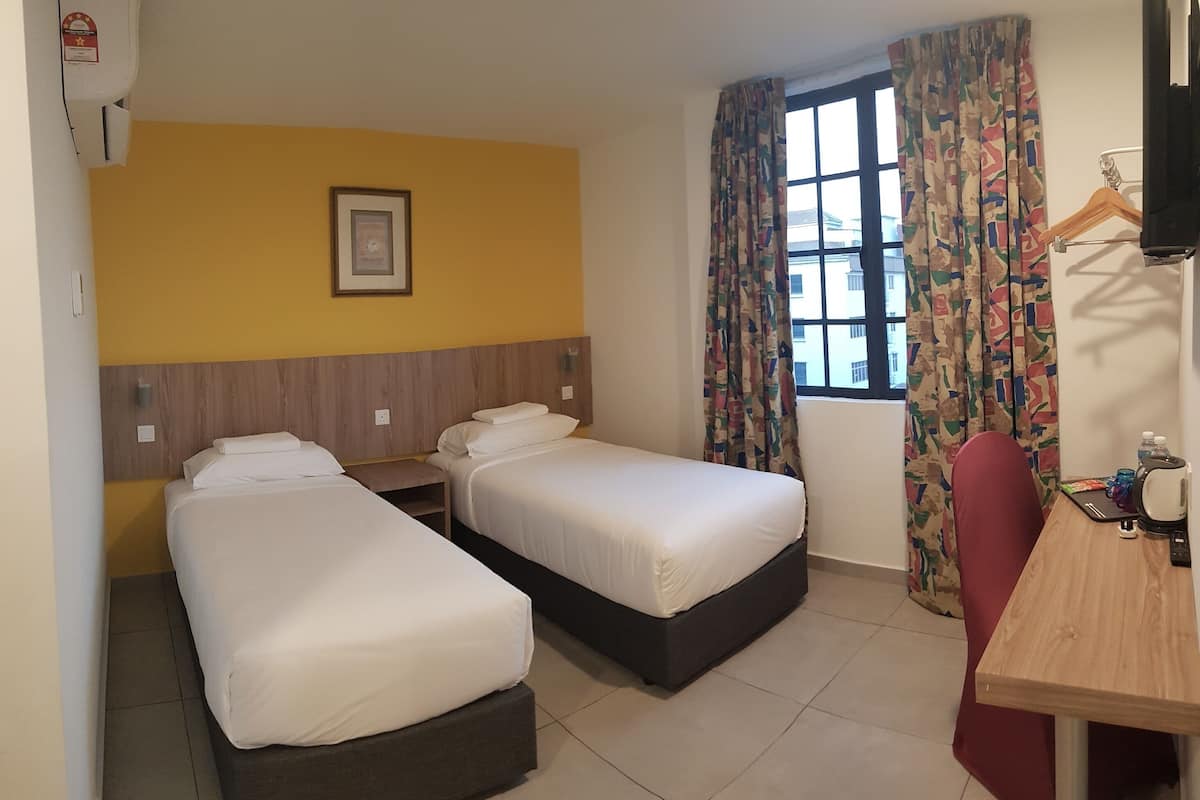 A hotel room with two single beds, a wooden headboard, a framed picture on the wall, a window with patterned curtains, and a desk with a chair.