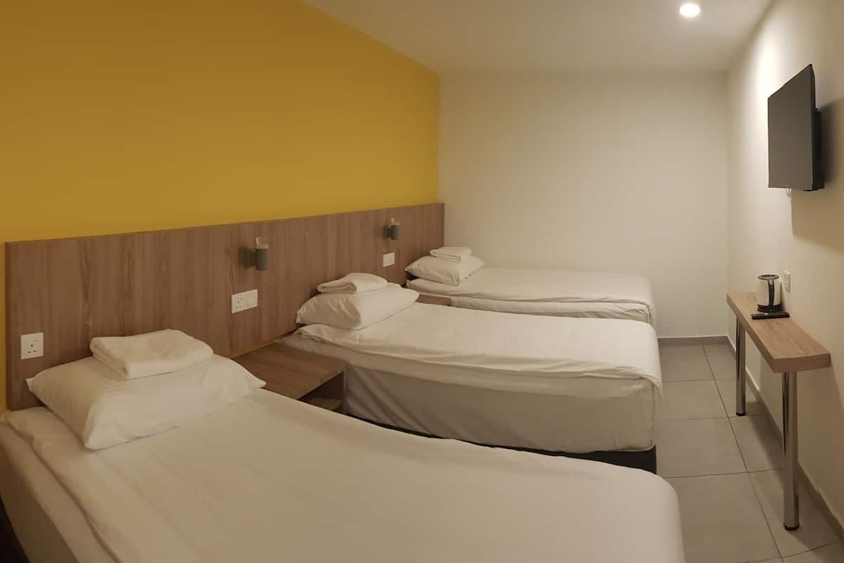 A hotel room with three beds, a wooden headboard, a TV mounted on the wall, and a small table with a kettle.