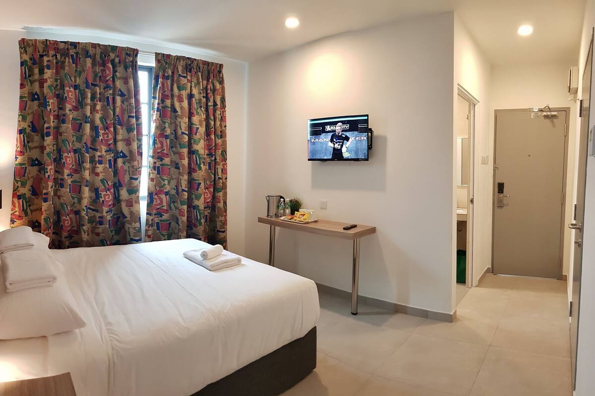 A hotel room with a bed, a bedside table with a TV, a small desk, and a door.