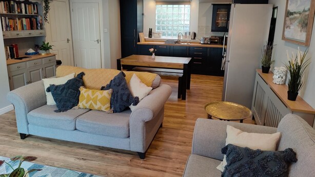 A modern living room with a grey sofa, a coffee table, and a small round table. There is a kitchen area with a window and a sink. - NEW Fabulous 2BD Maisonette Dunfermline, Fife (Dunfermline)