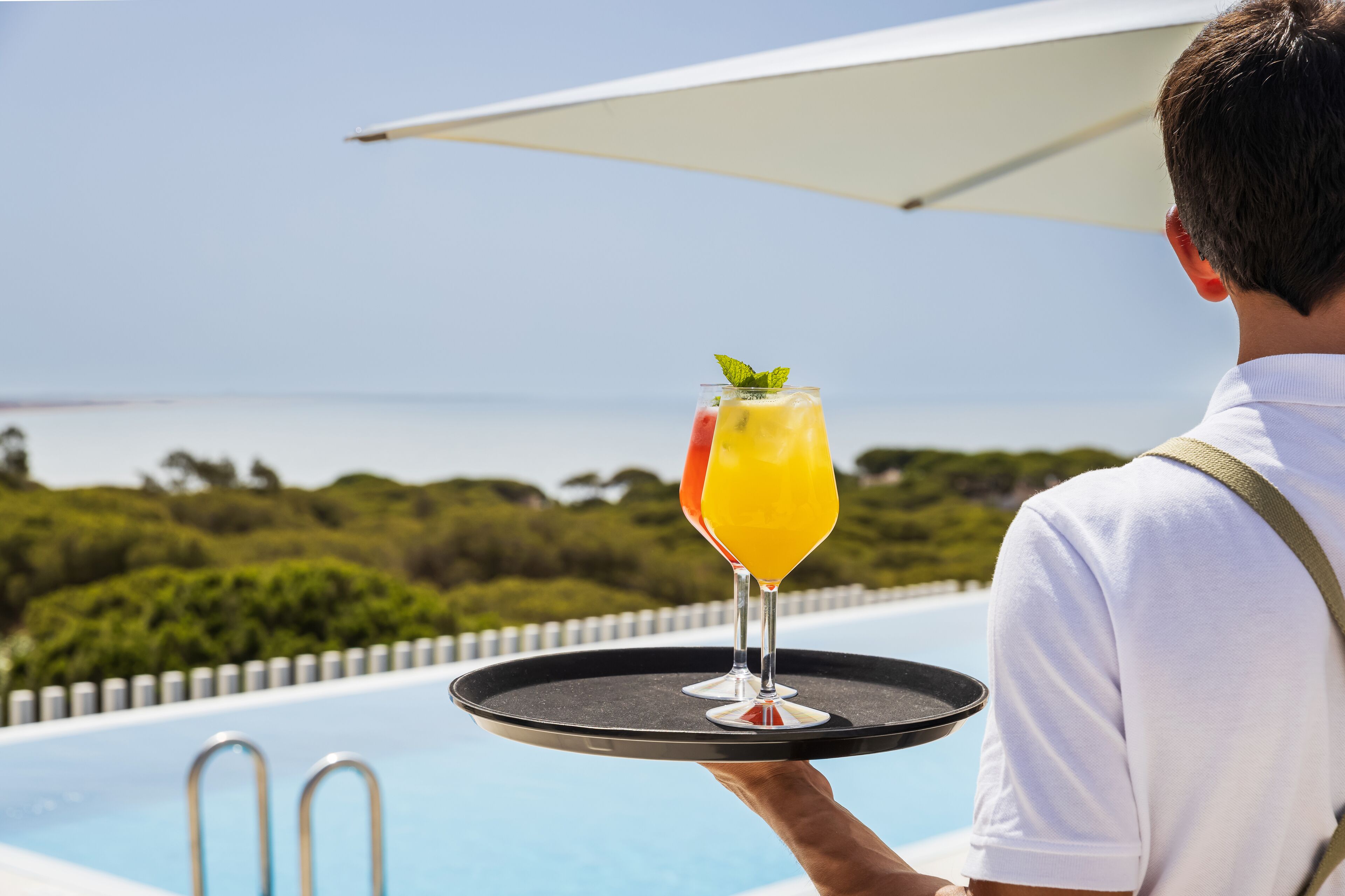 A person holding a tray with two drinks by a poolside with a view of the sea.