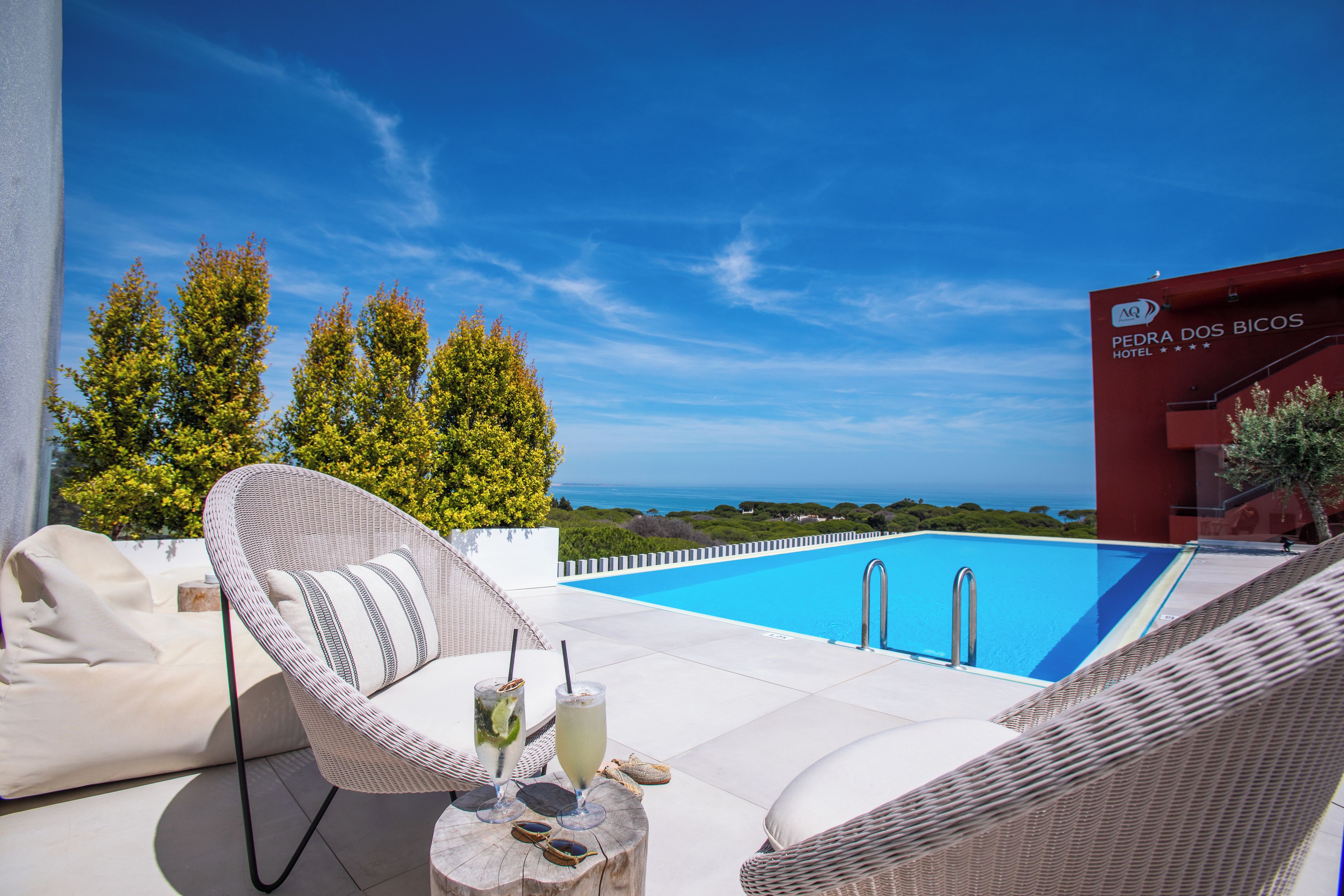 Zona de piscina con sillas de mimbre, una mesa con bebidas y vista al mar.