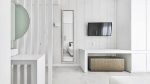 Una habitación moderna y minimalista con una paleta de colores blancos, un televisor de pantalla plana en la pared, un espejo redondo y un banco.
