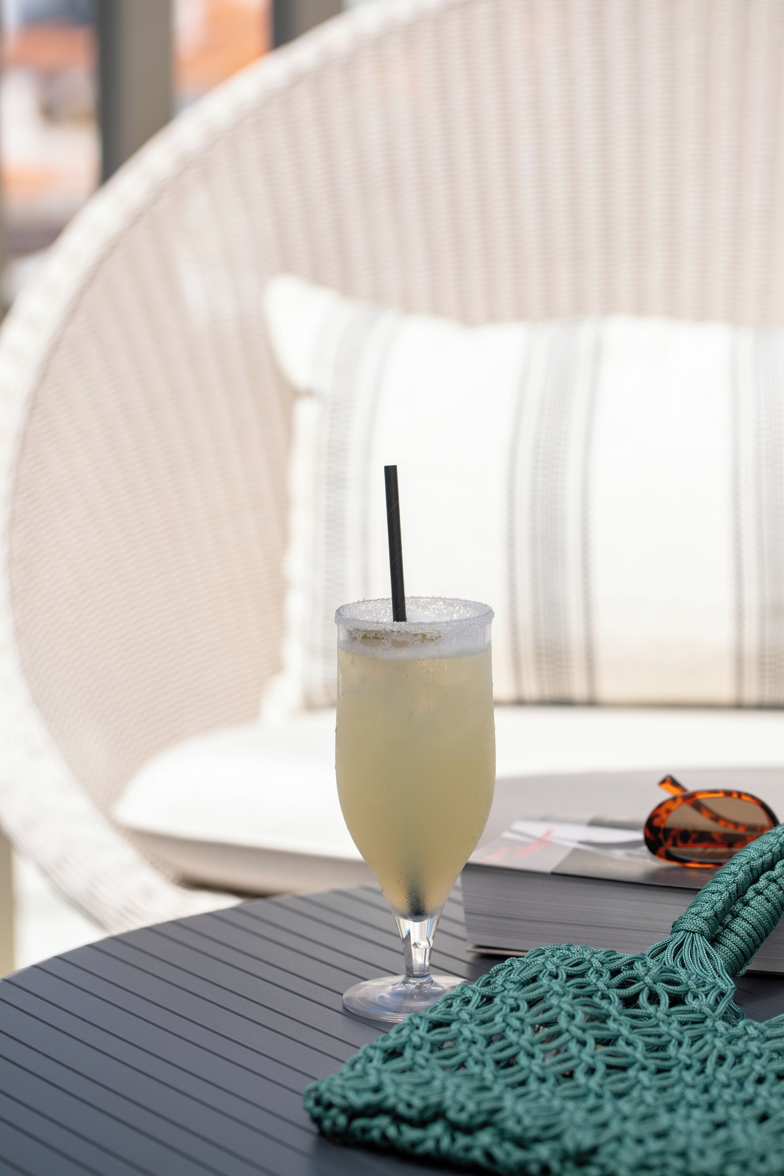 A glass of a light-colored beverage with a straw on a table, a green knitted item, and a white upholstered chair with a pillow in the background.