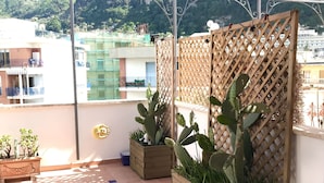 Ein Balkon mit Holz-Pflanzkästen, die Kakteen und andere Pflanzen beherbergen, einem Metallgeländer und Blick auf Gebäude sowie Grünflächen im Hintergrund.
