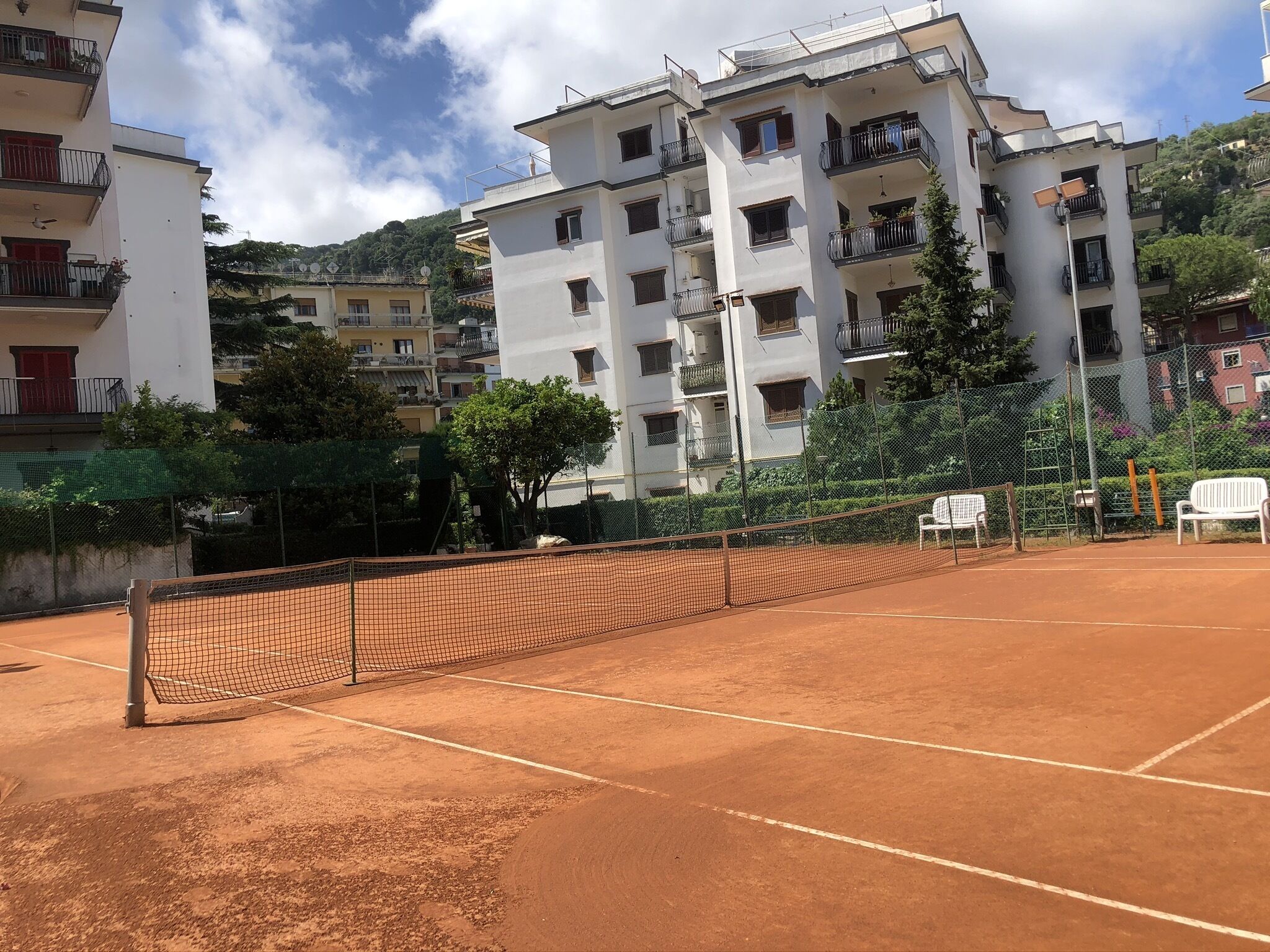 Ein Tennisplatz mit Netz, umgeben von Gebäuden und Grünflächen.