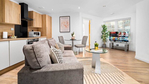 A modern living room with a grey sofa, wooden cabinets, a dining area with a glass table, and a TV displaying 'LIFE' magazine. - Luxury One Bed Apartment -  Jewellery Quarter (Birmingham)