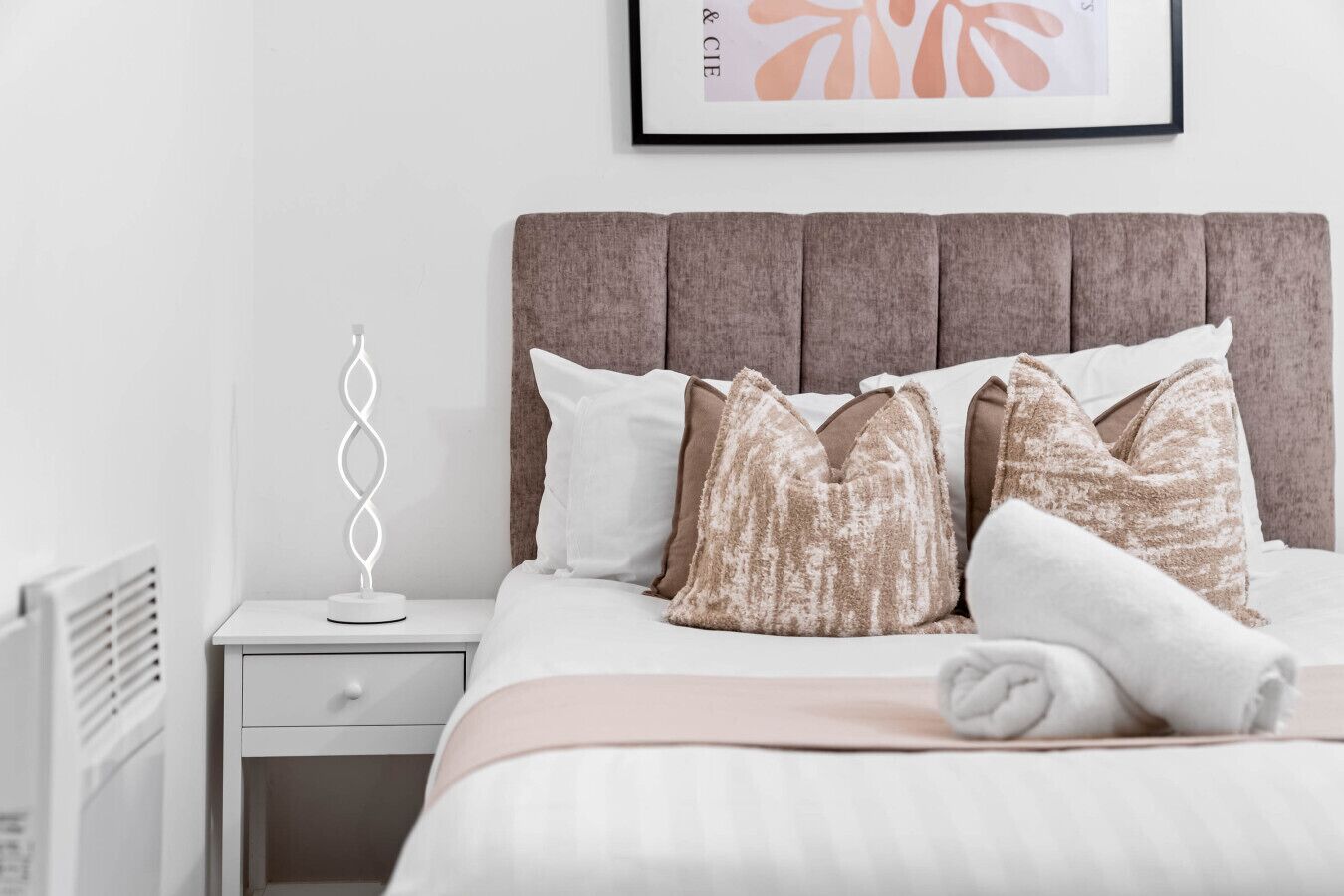 Una cama bien hecha con cabecera, una mesita de noche con lámpara y un cuadro enmarcado en la pared.