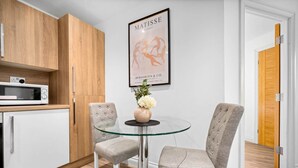 A modern kitchen with wooden cabinets, a microwave, and a glass table with two chairs. A framed artwork of a dancer is on the wall. - Luxury One Bed Apartment -  Jewellery Quarter (Birmingham)