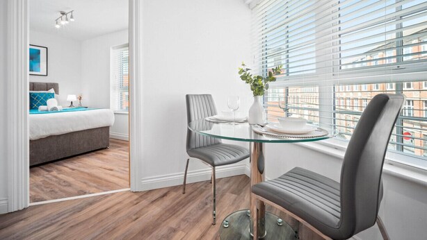 Een moderne hotelkamer met een glazen eettafel, grijs gestoffeerde stoelen, een kleine potplant en uitzicht op de stad door grote ramen.