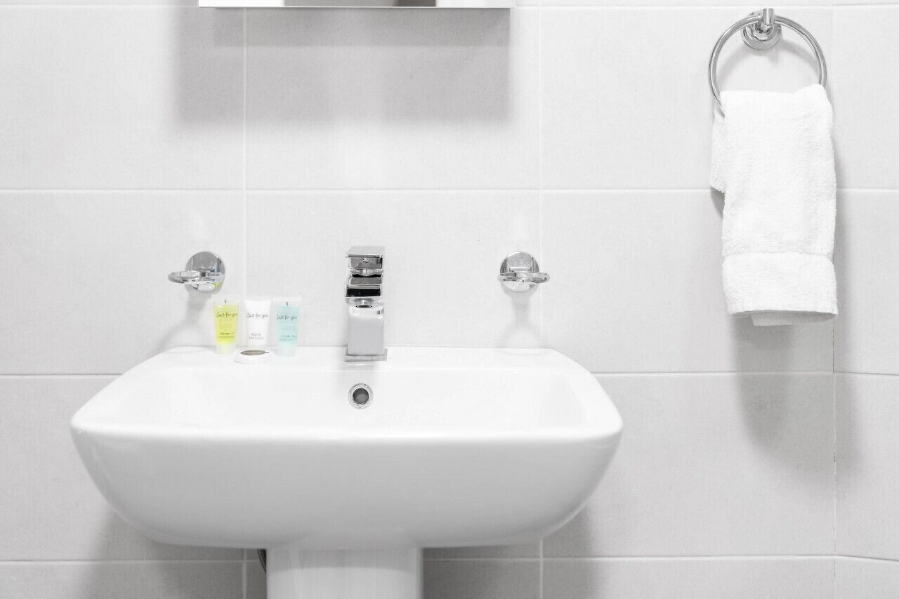 Un lavabo blanc dans une salle de bain, doté de deux robinets, avec un miroir au-dessus et une serviette suspendue à un anneau métallique.