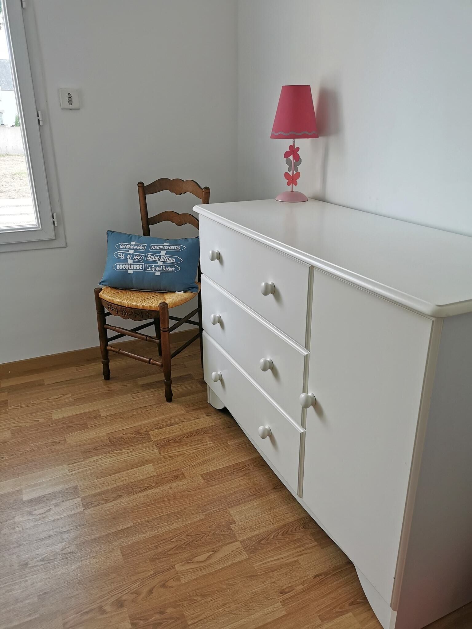A room with a wooden chair, a white dresser with drawers, a pink lamp, and a blue pillow with text.