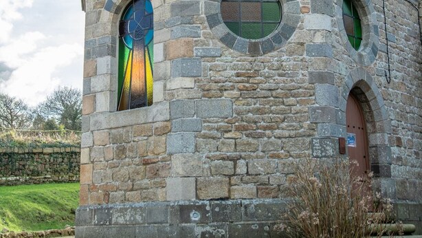A stone chapel with a cross on the roof, a stained glass window, and a small door.
