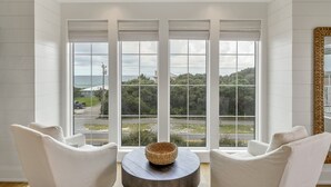 A room with a large window overlooking a road and greenery, two white armchairs, and a round wooden table with a woven basket.