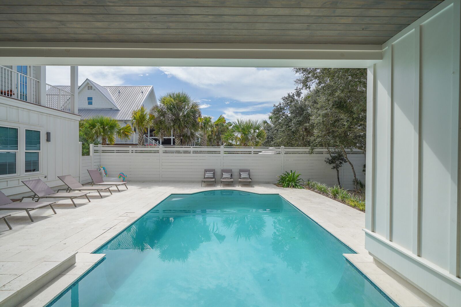 A modern outdoor swimming pool with lounge chairs, a covered patio, and a view of neighboring houses and trees.