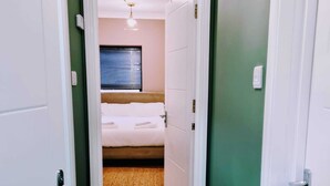 A narrow hallway leading to a bedroom with a bed, a window with blinds, and a wooden floor. - Beautiful & Clean 1 bed Apartment in Hackney Wick (London)