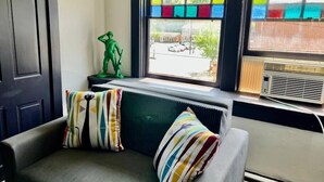 Ein Eckzimmer mit grauem Sofa, bunten Kissen mit Streifen, einer grünen Statue, einem Heizkörper und einem Fenster mit farbigem Buntglasbild.