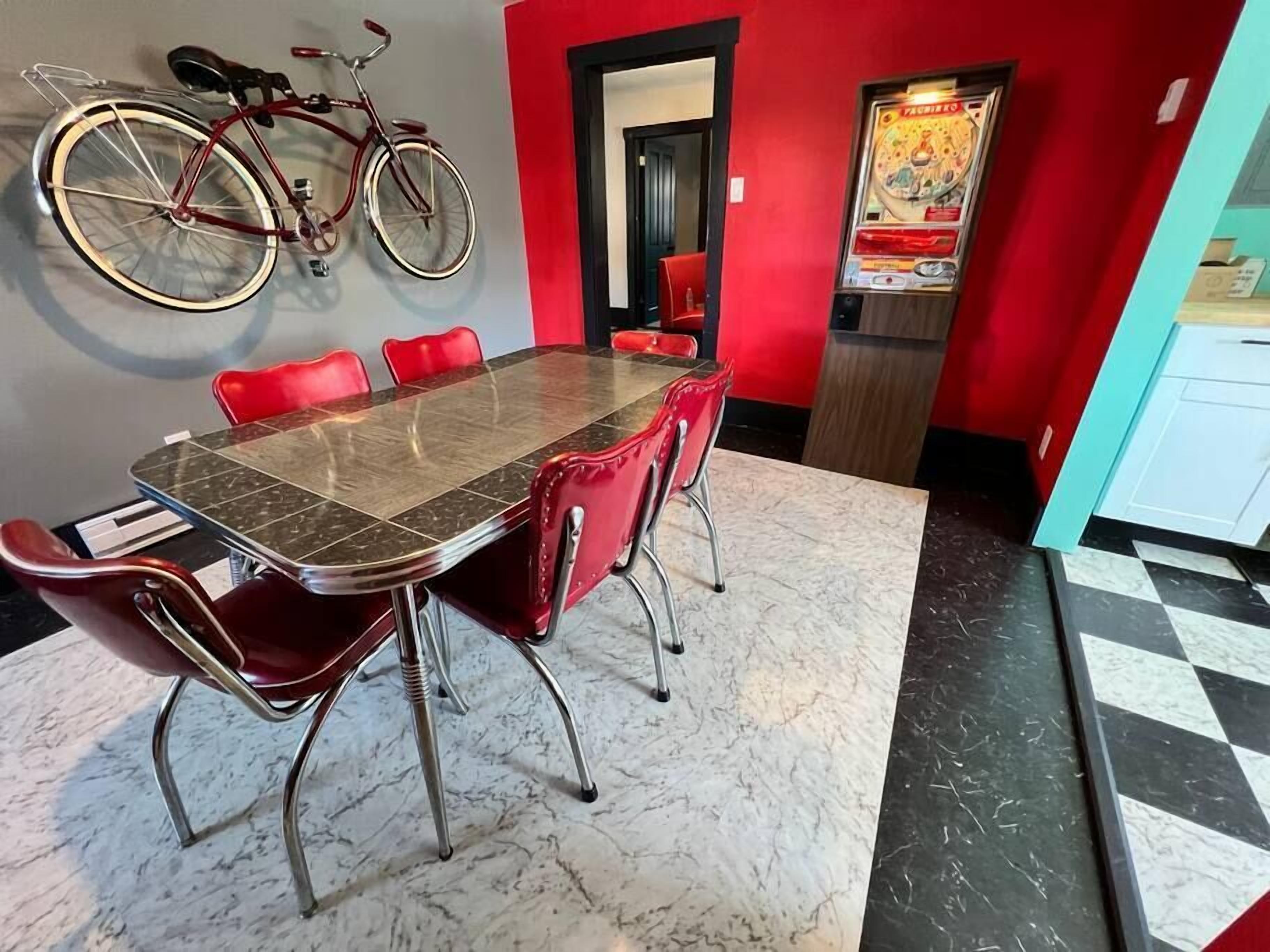 Un comedor con una mesa de mármol y sillas rojas, una bicicleta colgada en la pared y una máquina recreativa de pizza.