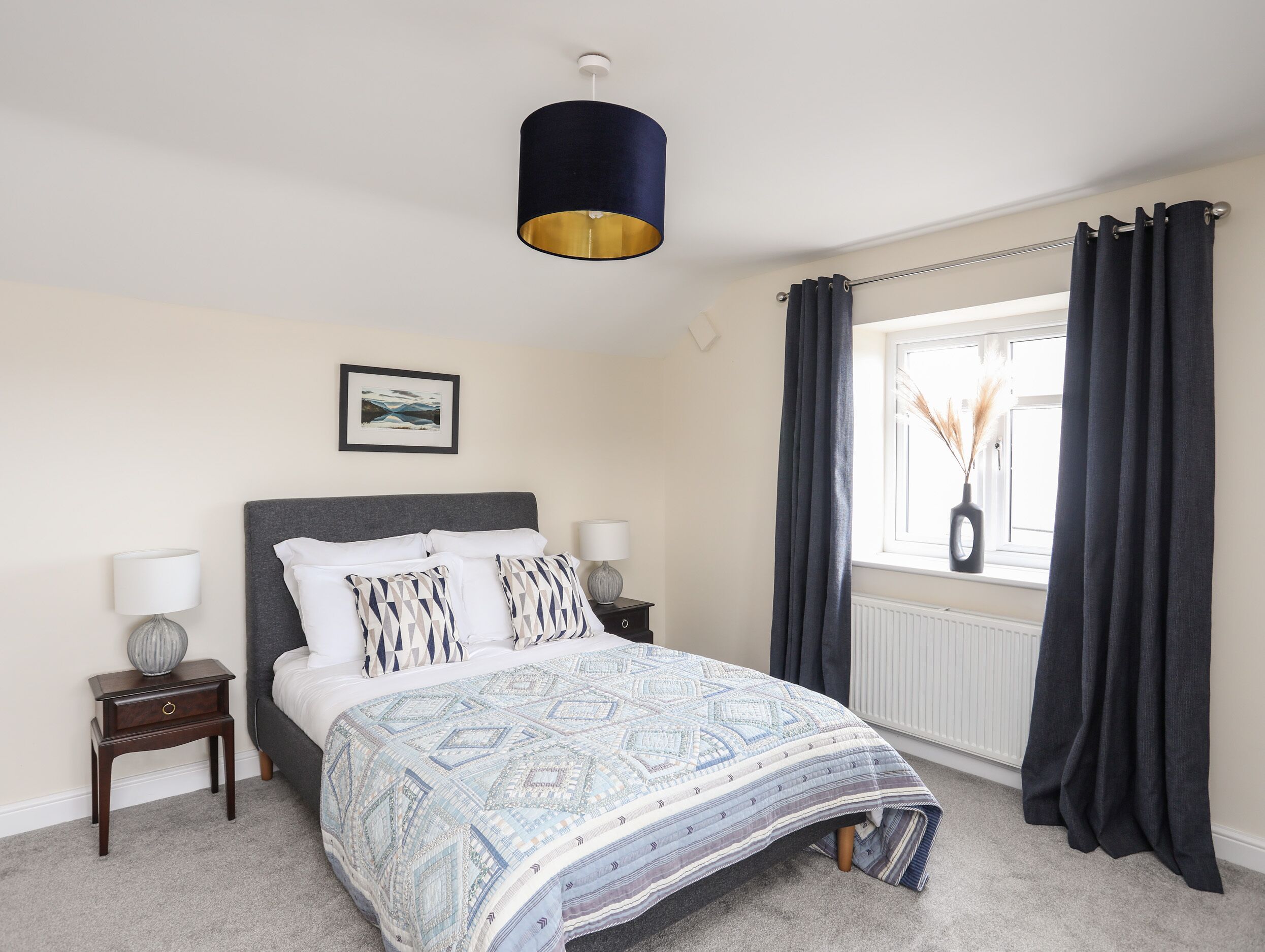 A bedroom with a bed, bedside tables, a hanging light fixture, a window with curtains, and a wall-mounted picture.
