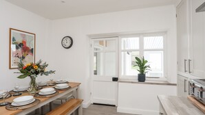 A modern kitchen with white cabinetry, a dining area with a wooden table set for four, and a potted plant on the windowsill. - Gwynant - Bull Bay (Amlwch)