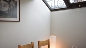 Un comedor con una mesa dispuesta para dos personas, que incluye platos, tazones, cubiertos y vasos. Hay una lámpara sobre la mesa y un cuadro enmarcado en la pared.