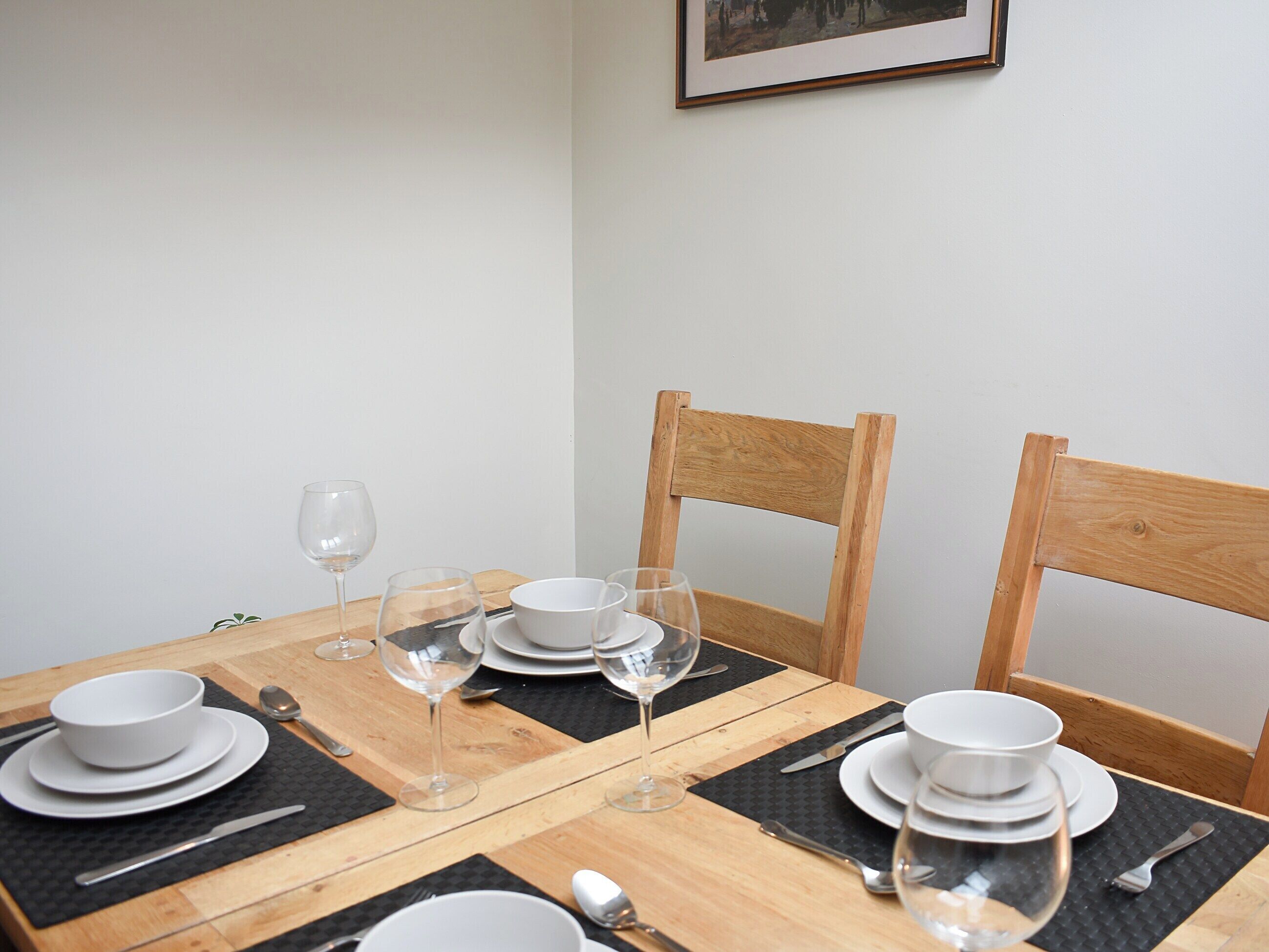 A dining table set with plates, bowls, cutlery, and wine glasses, with wooden chairs and a framed picture on the wall.