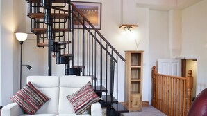 Una sala de estar con sofá blanco, almohadones a rayas, una mesa de centro de madera, una escalera espiral negra, una estantería de madera y una puerta blanca.