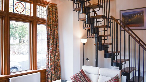 Una habitación con un sofá blanco, una mesa de madera, un radiador, una ventana con cortinas y una escalera espiral negra.