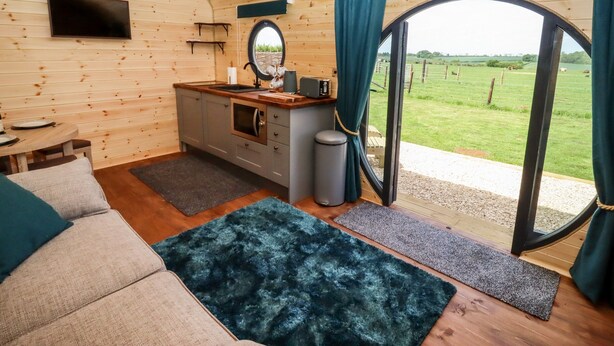 A cozy cabin interior with a wooden interior, a kitchenette, a sofa, and a large window offering a view of a green field. - The Happy Valley Pod (Chipping Norton)