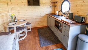 A cozy, wooden interior with a round dining table, two chairs, a small kitchen area with a sink and a microwave, and a window. - The Happy Valley Pod (Chipping Norton)