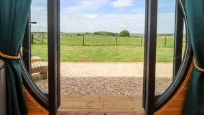 A view from inside a room with a large window, looking out to a grassy field with a fence and trees in the distance. - The Happy Valley Pod (Chipping Norton)