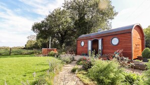 A wooden cabin with a circular window and a door, surrounded by a garden with various plants and a gravel path. - The Happy Valley Pod (Chipping Norton)
