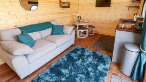 A cozy cabin interior with a sofa, a small table with a vase of flowers, a TV mounted on the wall, and a trash can. - The Happy Valley Pod (Chipping Norton)