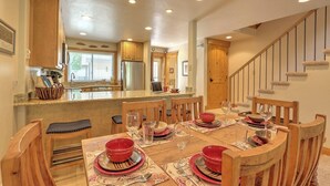On aperçoit un coin repas avec une table en bois dressée pour quatre personnes, entourée de chaises. Au fond, on distingue une cuisine avec un réfrigérateur et une fenêtre.