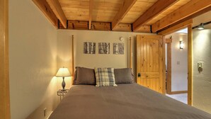 Une chambre avec un plafond en bois, un lit avec une literie marron, une table de chevet avec une lampe et une porte donnant sur une autre pièce.