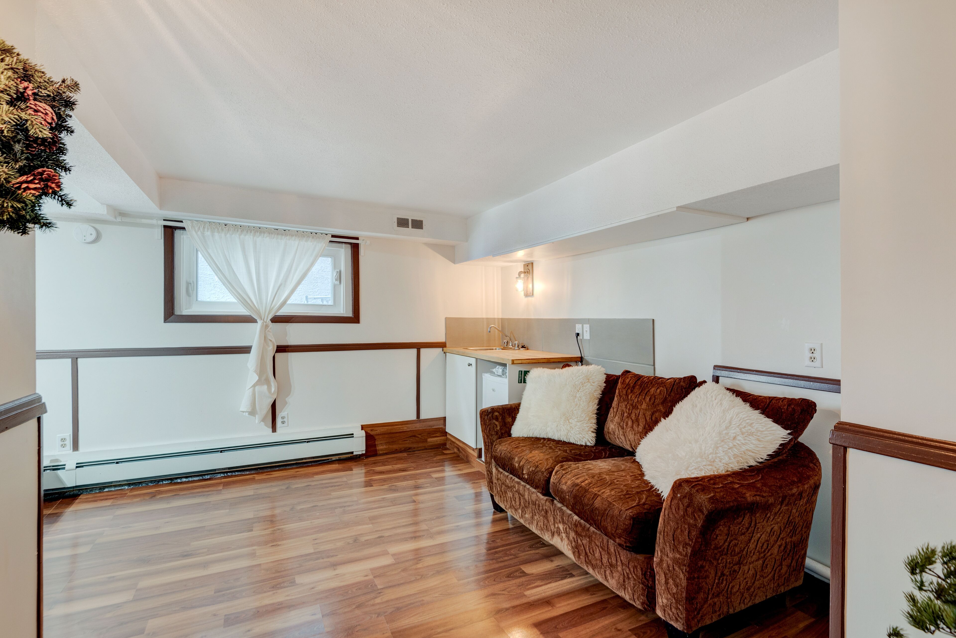 A cozy living room with a brown sofa, a small kitchen area, and a window with white curtains.