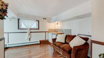 A cozy living room with a brown sofa, a small kitchen area, and a window with white curtains.