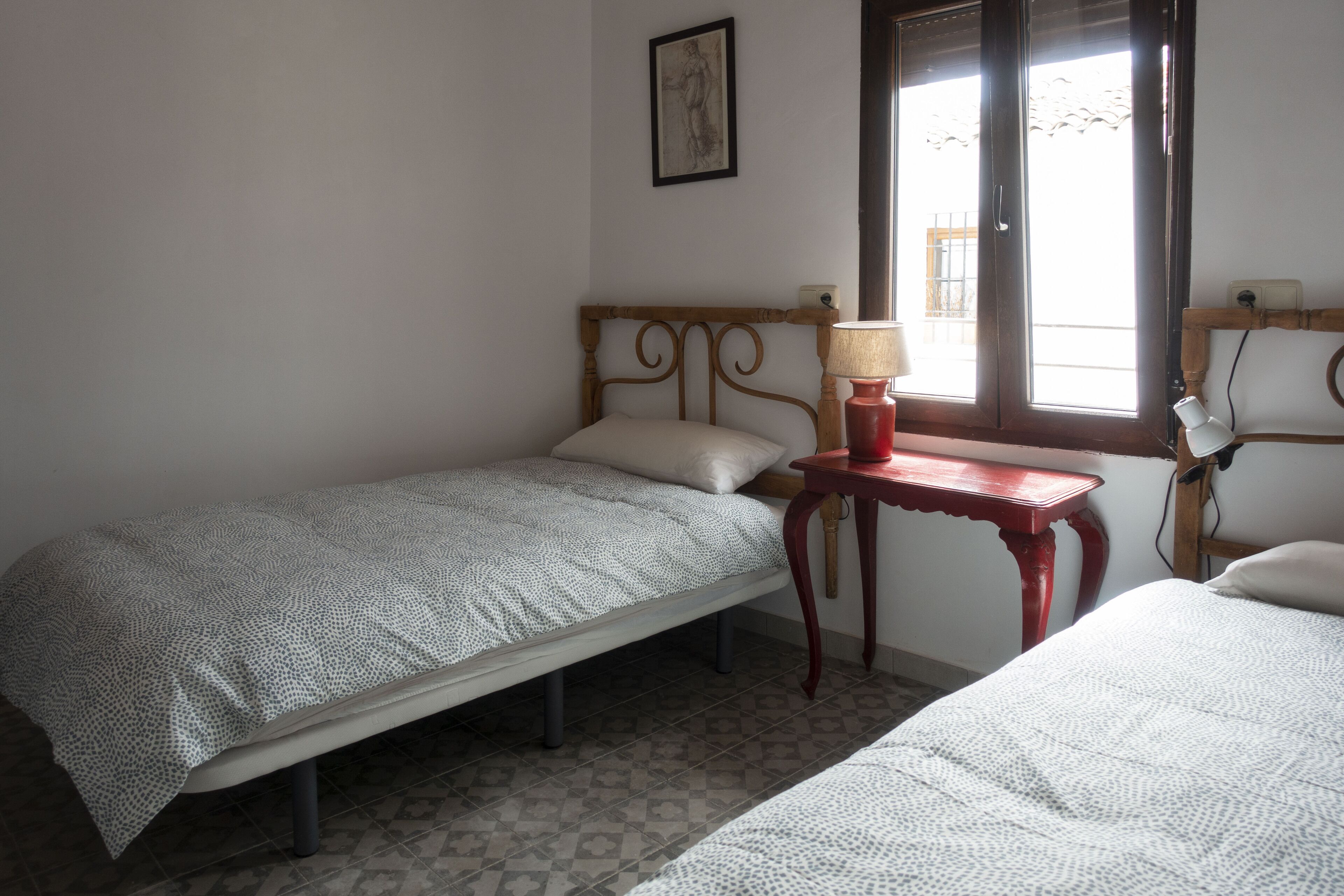 Un dormitorio con dos camas individuales, una mesita de noche con lámpara, una ventana con vistas y un interruptor de luz en la pared.
