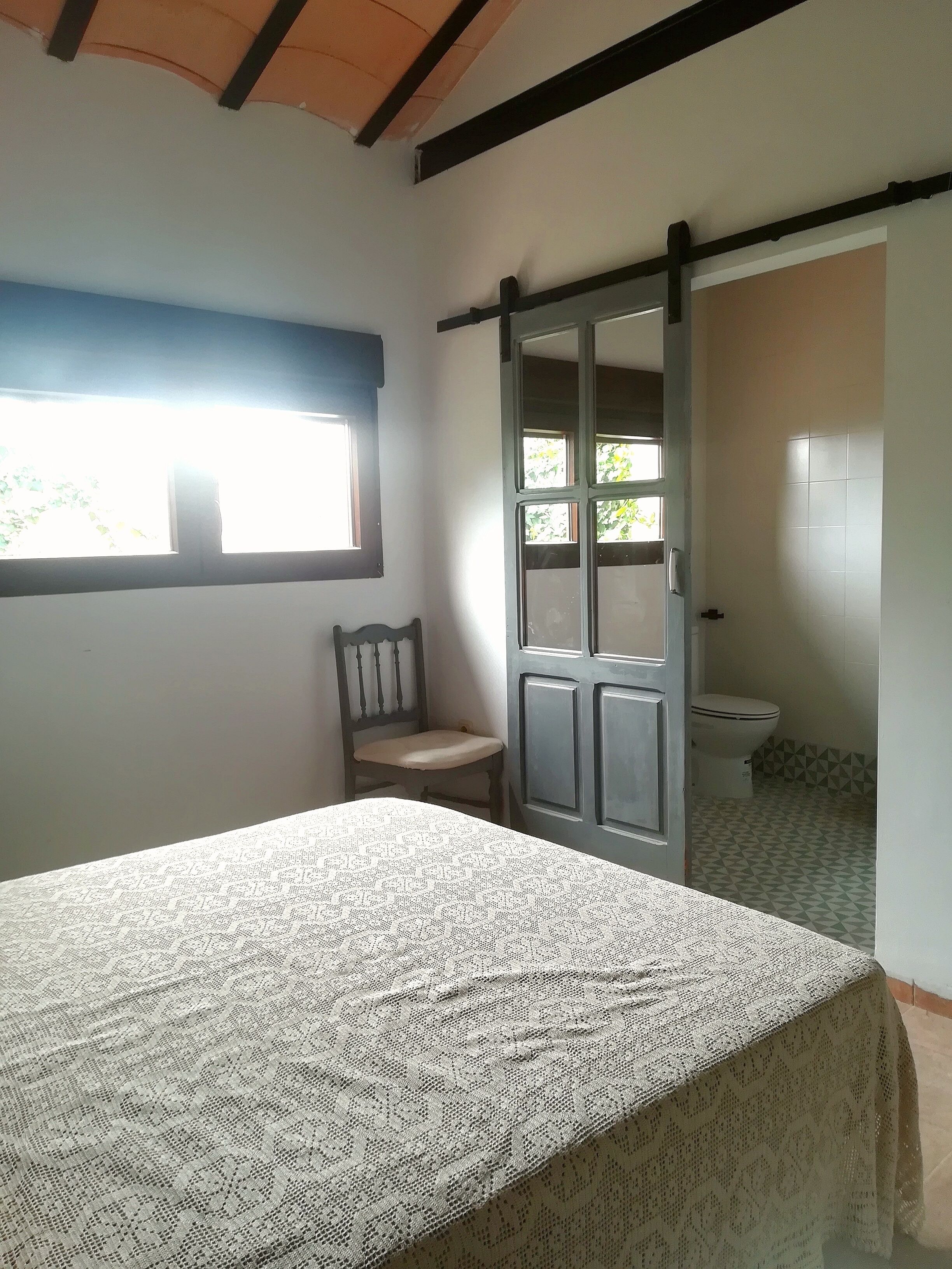 Un dormitorio con cama, una silla, una ventana y un baño visible a través de una puerta abierta.
