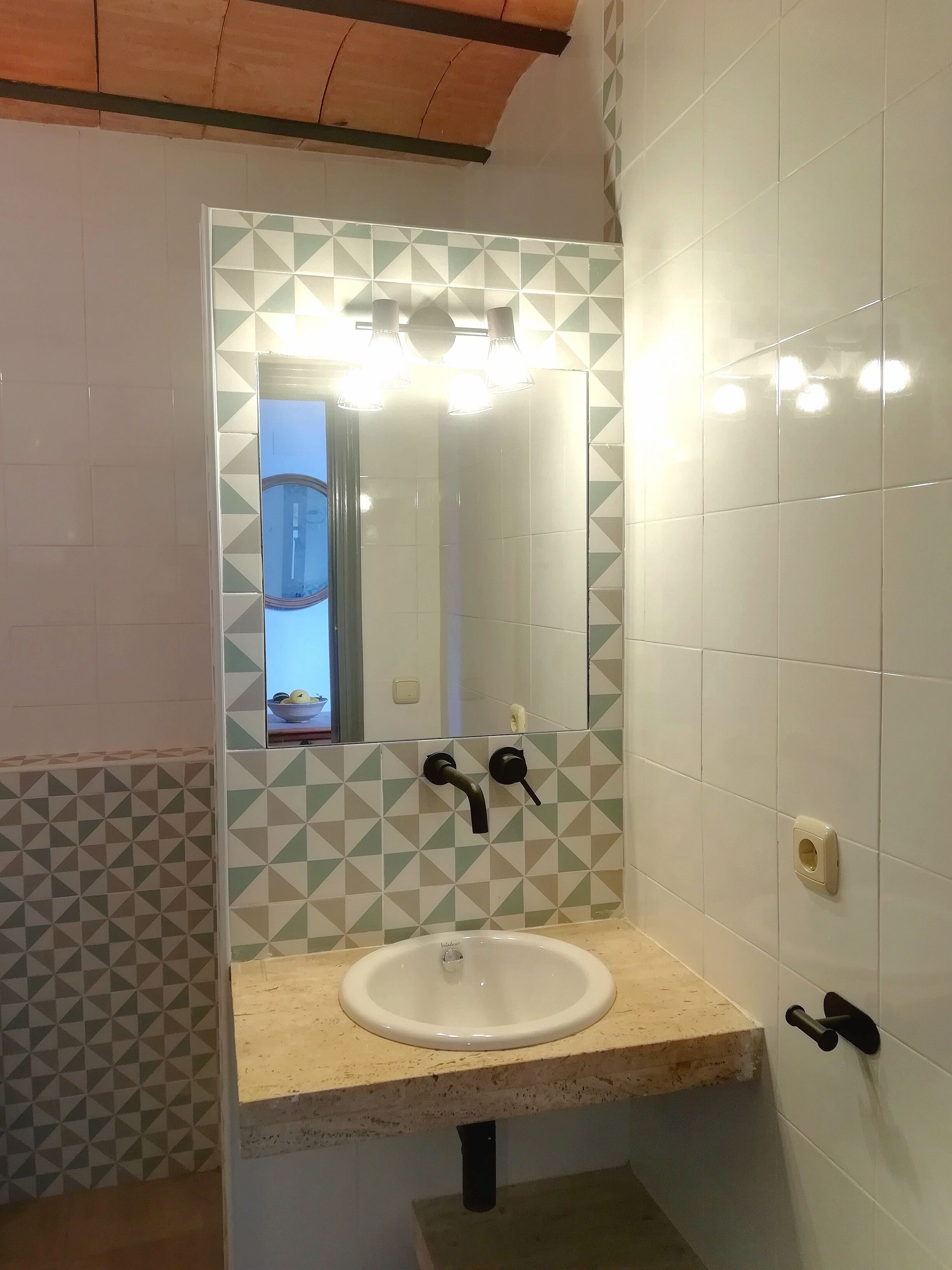 Ein Badezimmer mit einem weißen Waschbecken, zwei schwarzen Wasserhähnen, einem Spiegel und einem geometrischen Fliesenmuster an den Wänden.