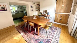 A dining area with a wooden table, chairs, a rug, and a kitchen in the background. - Widgeons Keel (Falmouth)
