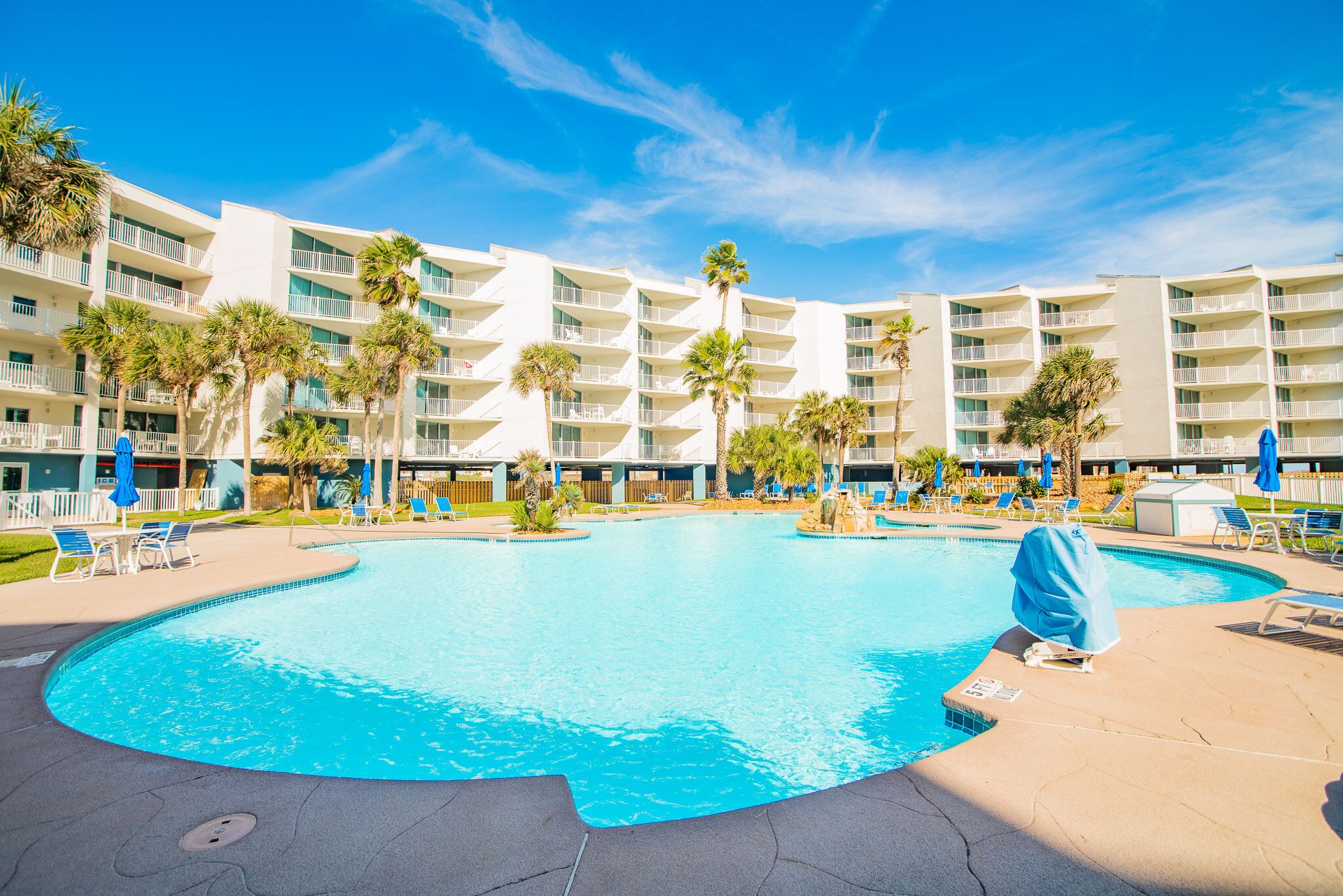 A large swimming pool surrounded by a hotel with multiple balconies and palm trees.
