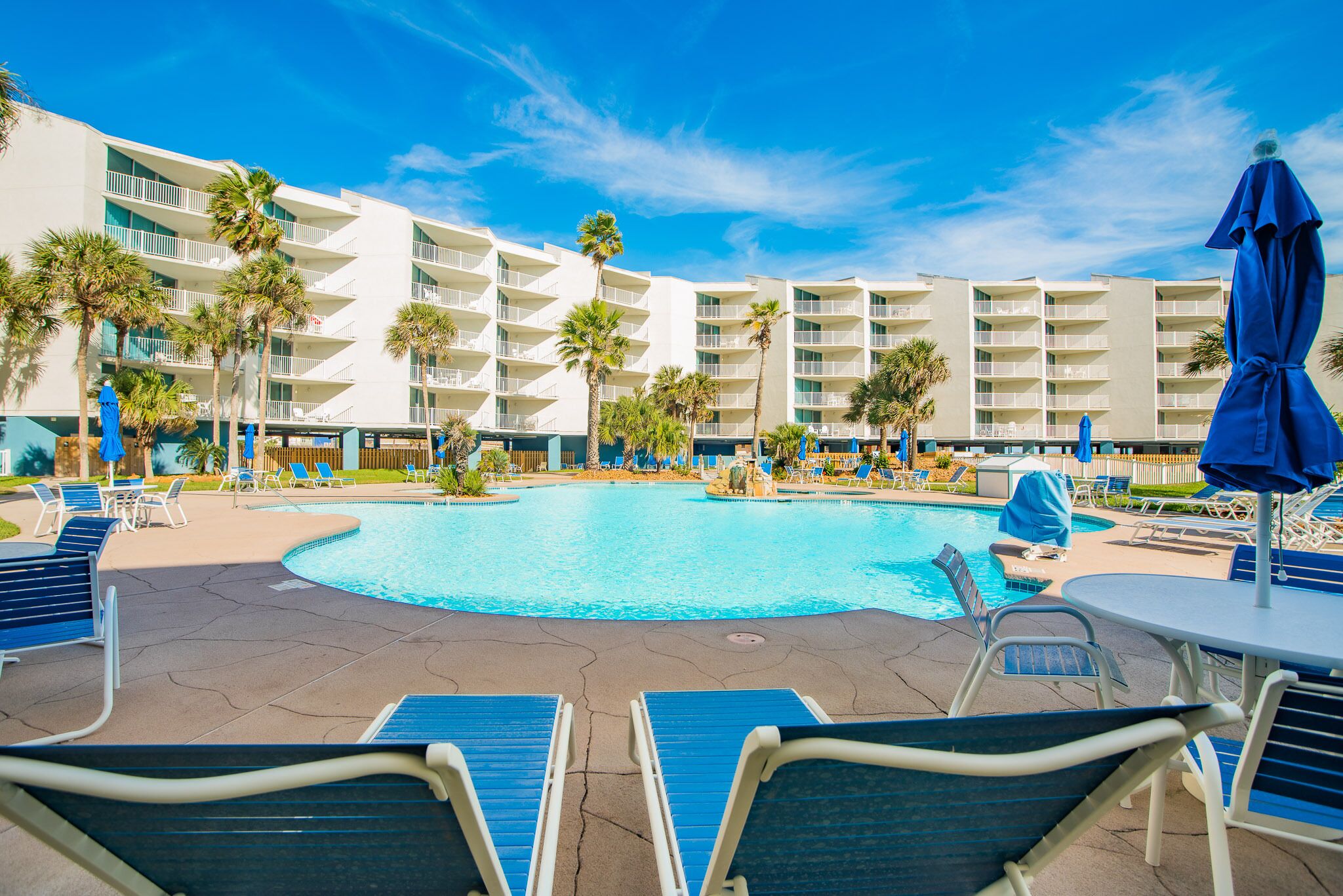 Ein Hotelpoolbereich mit mehreren Liegestühlen und Sonnenschirmen, umgeben von Palmen und einem großen Pool.