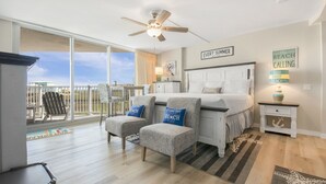A bedroom with a large bed, two armchairs, a ceiling fan, a balcony with a view, and a beach-themed decor. - Sandcastle Condominium Unit 128 (Port Aransas)
